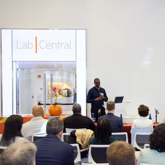 A biotech entrepreneur stands at a podium in LabCentral's lobby, delivering a Golden Ticket pitch to an audience of sponsors, investors and community members.